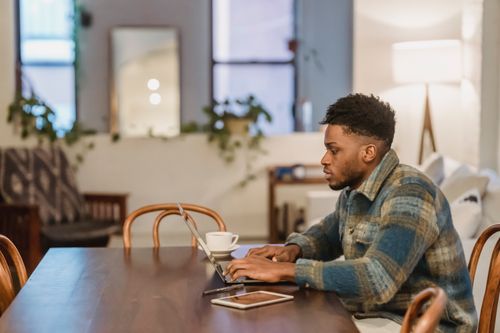 Free Side view of young bearded African American male in casual clothes and sitting at table with laptop and cup of coffee while working on project Stock Photo