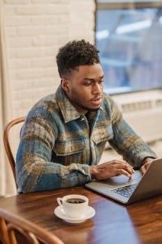 Young bearded African American man in casual clothes sitting at table with laptop and cup of coffee while working on remote project in home office