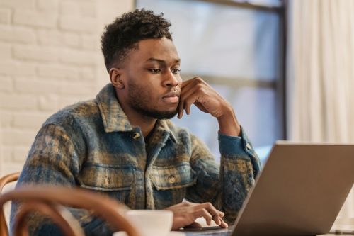 Free Concentrated bearded African American male freelancer in casual clothes sitting at table with laptop and leaning on hand while working on remote project in home workspace Stock Photo