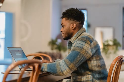 Free Side view of young bearded black male freelancer with curly hair in casual clothes and sitting at wooden table and browsing netbook while working on project at home Stock Photo