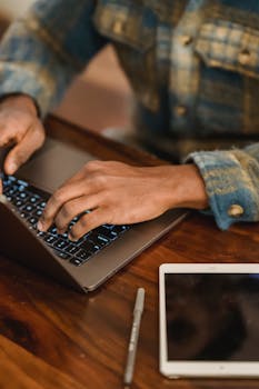 Crop faceless male freelancer sitting at wooden table with tablet and pen and using laptop while working remotely