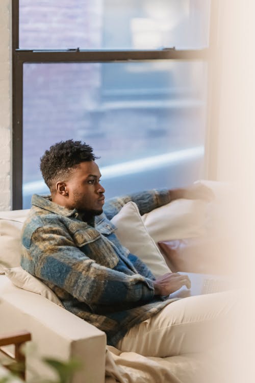 Side view of young African American male freelancer sitting on couch with laptop on knees and thoughtfully looking at screen