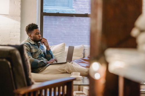 Free Side view of pensive young African American male freelancer looking at screen and touching chin while working remotely on laptop from comfortable sofa Stock Photo