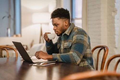 Free Focused bearded African American male in casual clothes sitting at dinner table with cup of tea and attentively looking at screen while browsing laptop Stock Photo