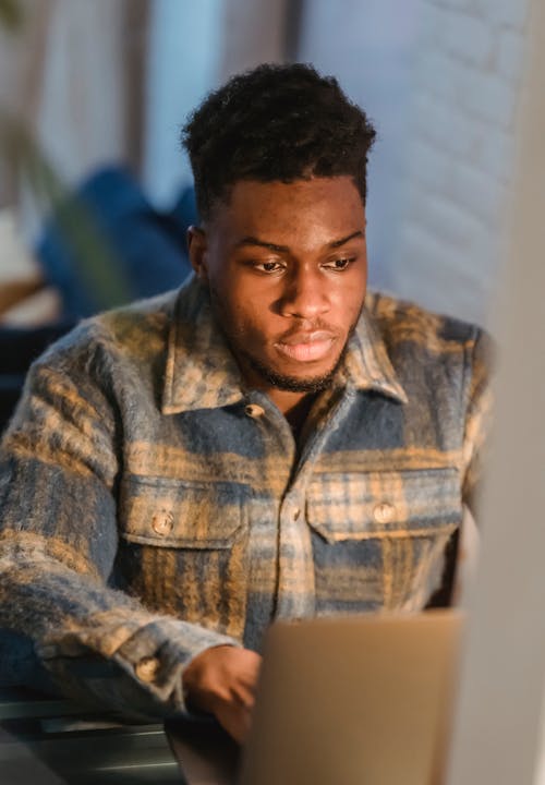 Focused African American young male in casual clothes looking at screen of laptop and typing on keyboard at home workplace
