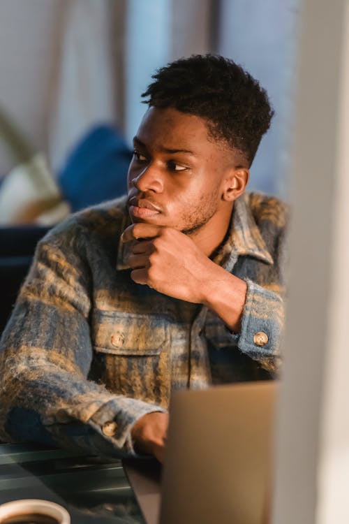 Serious young African American male freelancer thoughtfully looking away and touching chin while working on computer