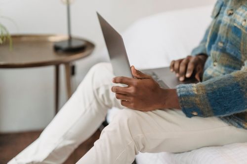 Free Side view of crop faceless African American male sitting on comfortable sofa and surfing internet on portable computer while remotely working at home Stock Photo