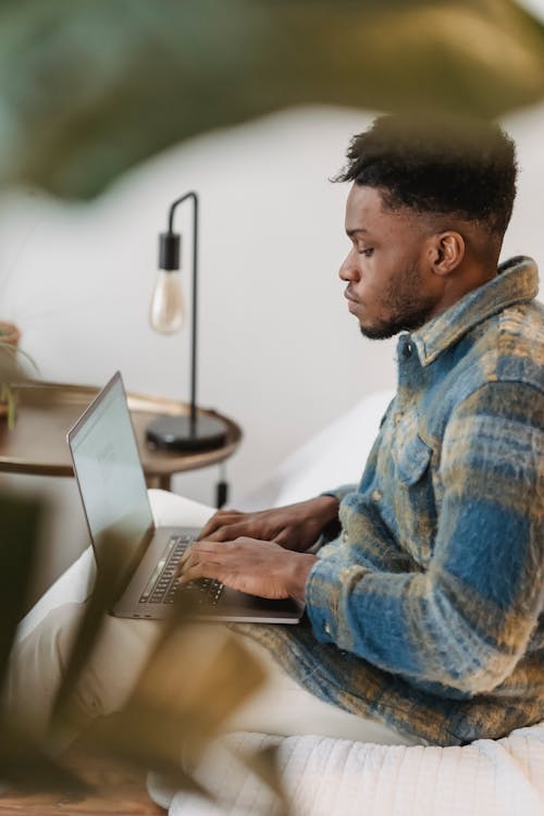 Side view of concentrated African American guy in stylish shirt sitting on comfortable sofa at home and typing on laptop while working remotely
