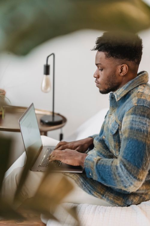 Free Side view of concentrated African American guy in stylish shirt sitting on comfortable sofa at home and typing on laptop while working remotely Stock Photo