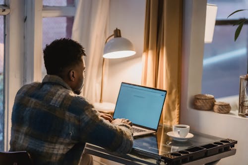 Free Back view of concentrated African American male typing on keyboard of laptop while sitting at table with cup of coffee and light from lamp Stock Photo