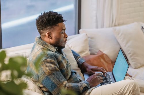 Free Side view of concentrated African American male sitting on soft couch near window and browsing internet on laptop and looking at screen Stock Photo