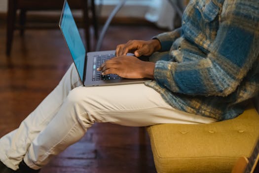 Side view of crop unrecognizable African American male freelancer sitting on yellow banquet and typing on keyboard