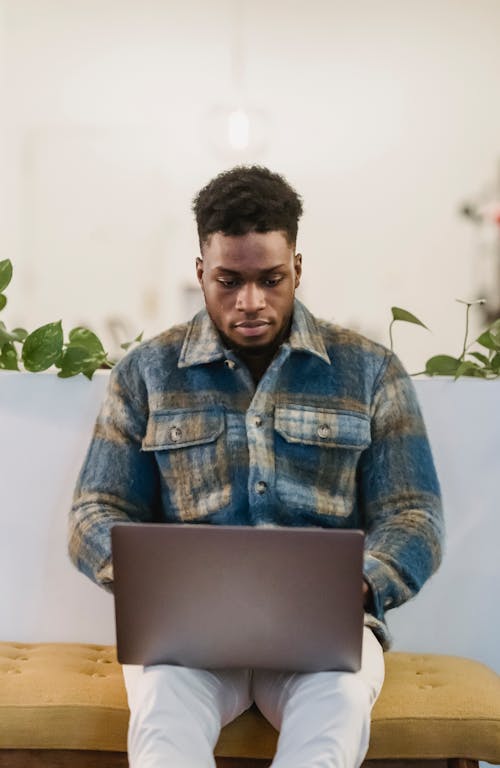 Concentrated young African American male freelancer in checkered shirt typing on laptop keyboard while working remotely in cafe