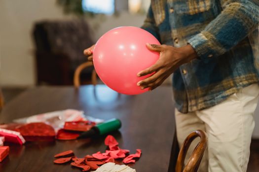Side view of crop unrecognizable African American male in casual clothes blowing balloons while preparing for party at home