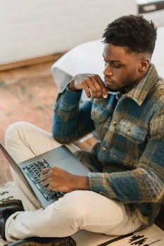 From above of concentrated young self employed ethnic man with dark Afro hair in casual clothes sitting on floor and working remotely on laptop at home
