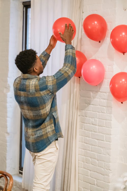 African American male in casual clothes decorating white wall of home with red and pink balloons preparing to Saint Valentine day