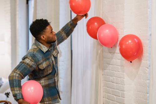 Free Side view of cheerful young African American male in casual clothes with red and pink balloons standing near curtain in apartment Stock Photo