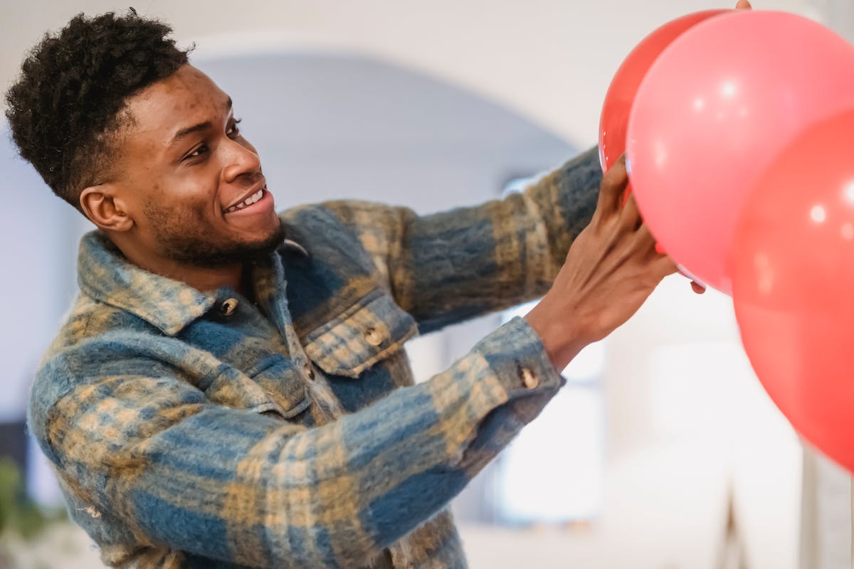 Bearded black man in checkered shirt correcting red balloons position while decorating room to special event