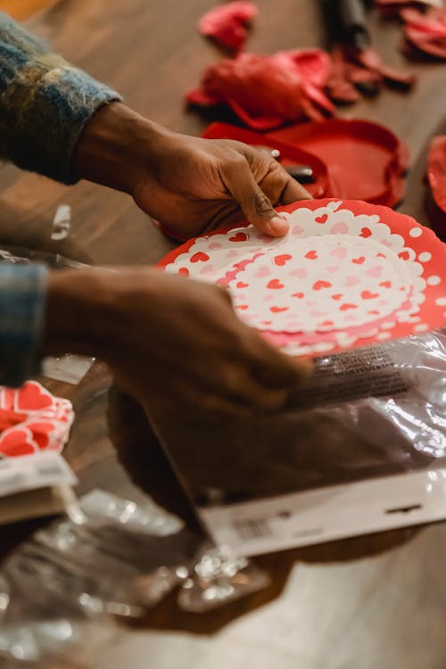 From above of crop anonymous African American male looking over festive napkins with red hearts on wooden table