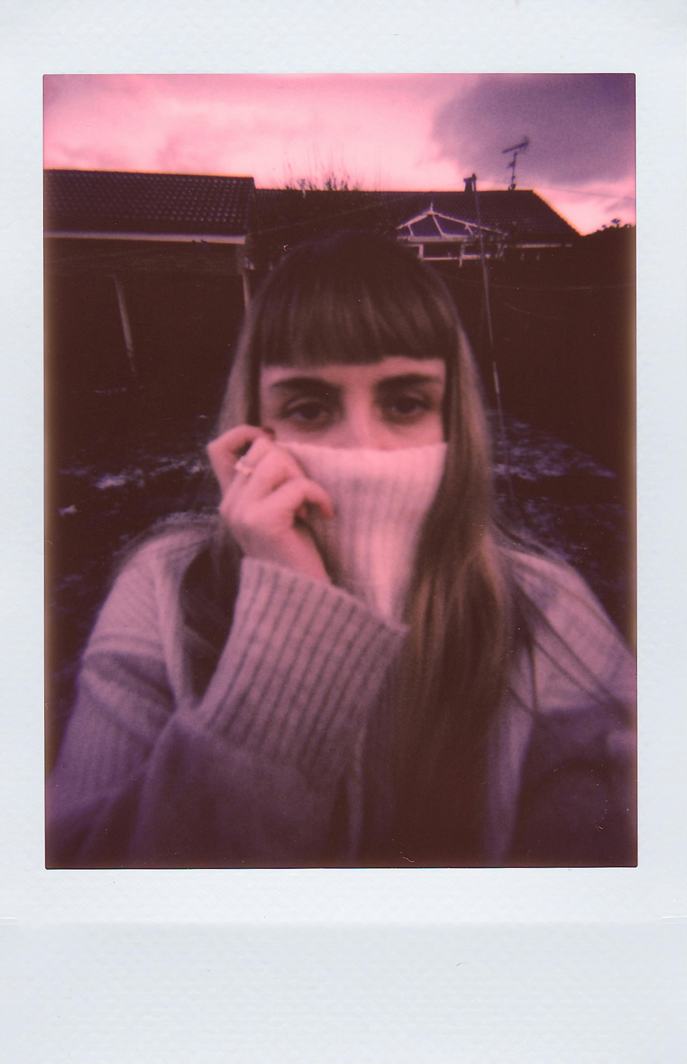 A young woman in a turtleneck sweater hides her face, set against a moody purple sunset sky.