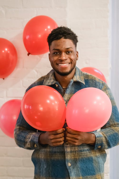 Portrait of smiling bearded African American guy in checkered shirt showing red balloons and looking at camera