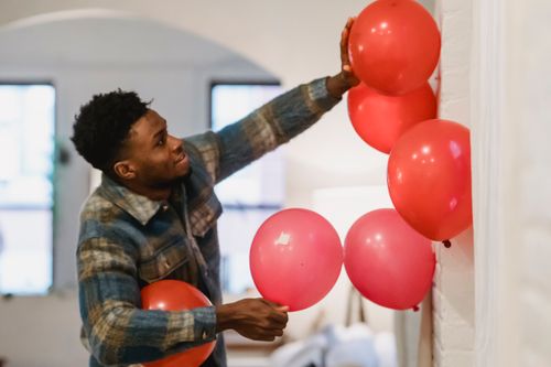 Free Side view of smiling African American male in casual shirt placing red balloons on white wall Stock Photo