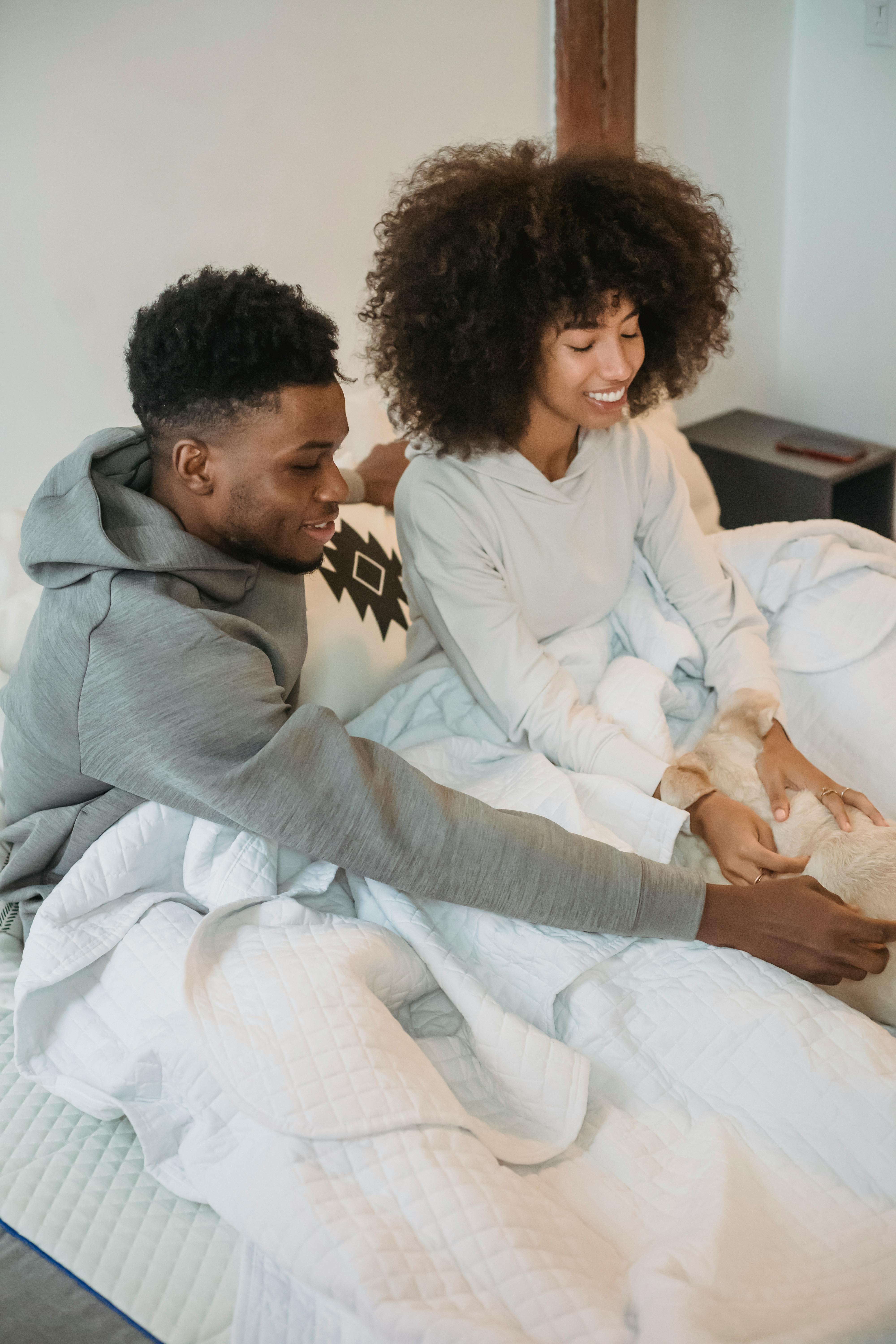 Happy young African American couple sitting on white bed and playing with dog