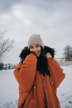 Happy young woman wearing warm jacket and hat while standing on snowy meadow near trees under cloudy gray sky in winter day and looking at camera