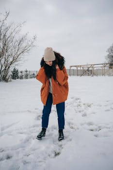 Full length of young woman wearing warm jacket and hat while standing on snowy meadow under gray cloudy sky in winter day near trees