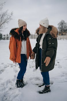 Two young women wearing winter coats and hats standing on a snowy field, chatting.