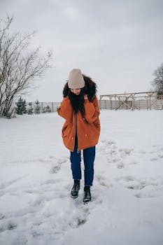 A young woman in a warm jacket and beanie stands in a snowy field, embracing winter's chill.