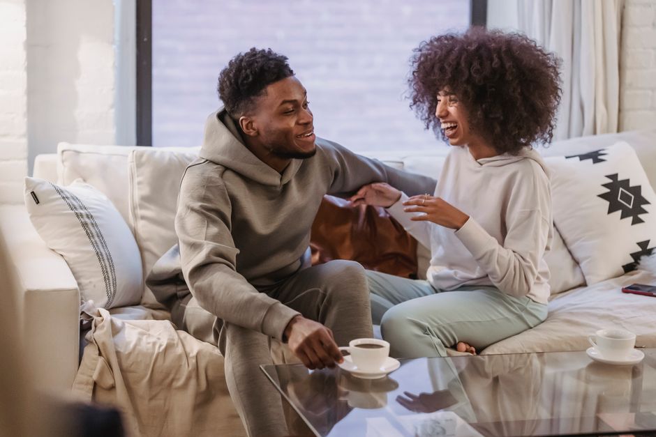 Positive African American couple talking and laughing on comfortable sofa at home in daytime