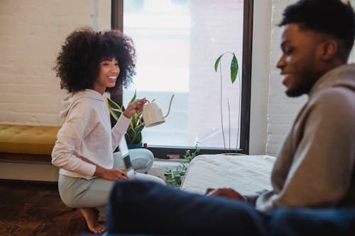 Smiling African American woman in casual clothes squatting down and watering plants while spending time with boyfriend browsing laptop at home in daytime