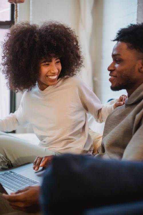 Free Smiling African American female laughing and looking at positive black boyfriend using laptop in light room in daytime Stock Photo