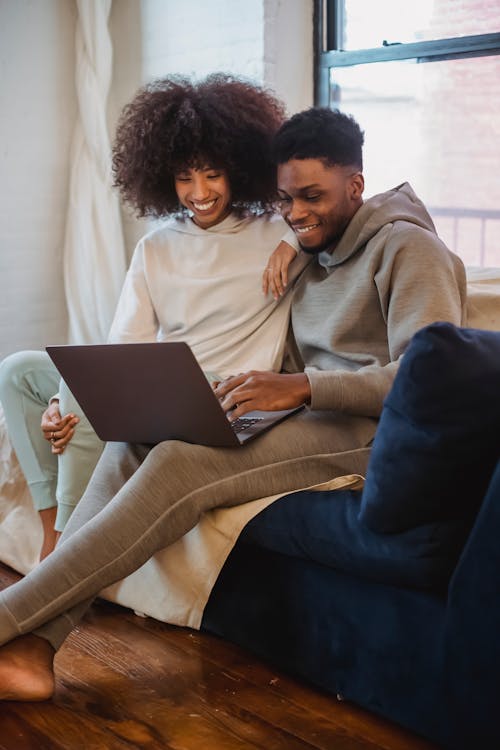 Happy African American couple sitting on comfortable sofa and watching video on laptop at home in daytime