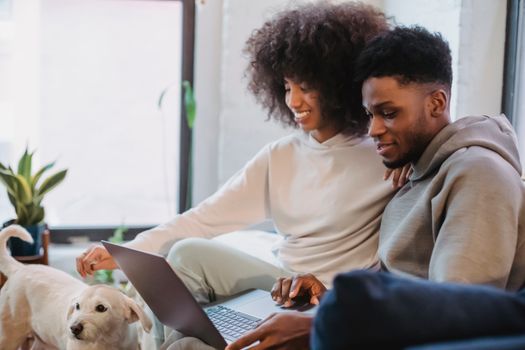 Positive African American female looking at calm dog and leaning on shoulder of black man browsing laptop at home in daytime