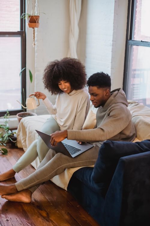 Free Smiling African American woman sitting with watering can on sofa and looking at laptop of boyfriend in living room in daytime Stock Photo