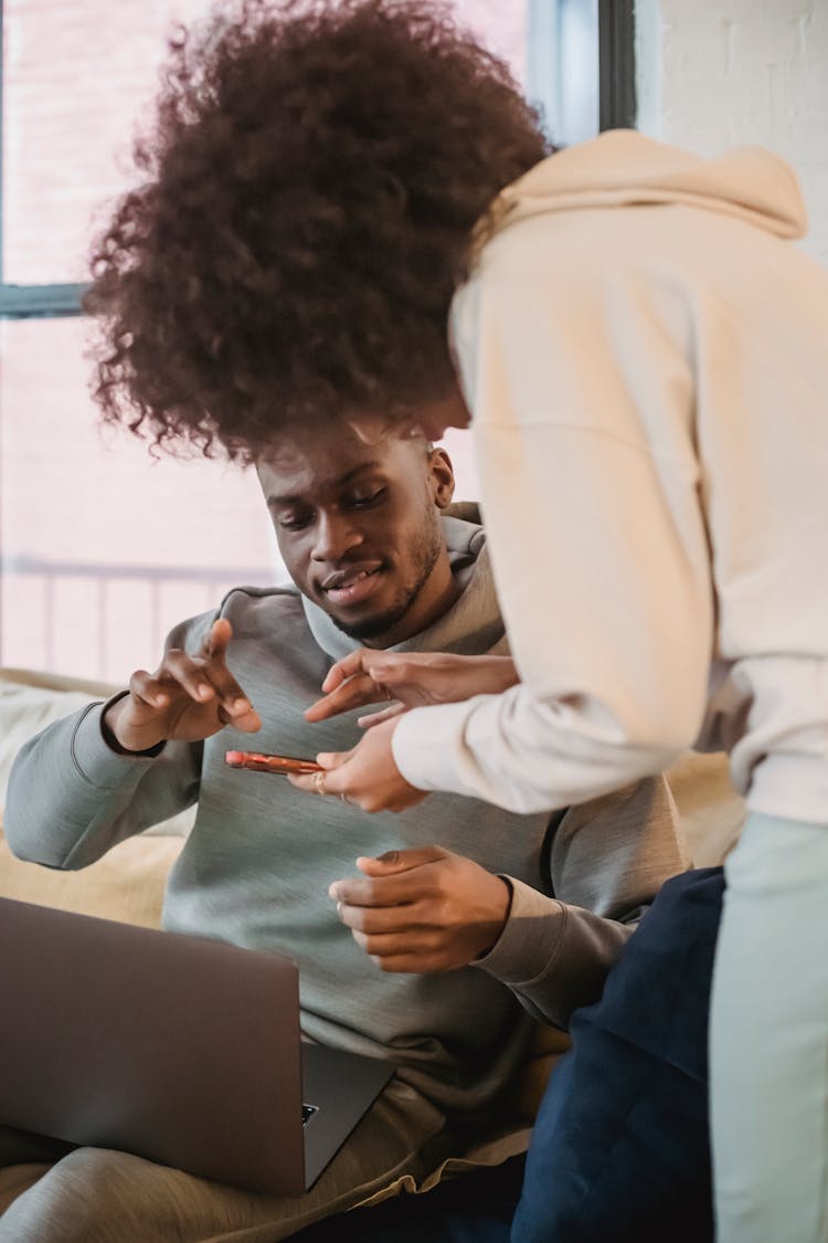 Black Female Showing Smartphone To Friend Sitting With Laptop