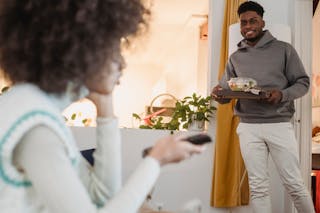 Smiling black man standing in room with bowl of salad near girlfriend sitting on couch