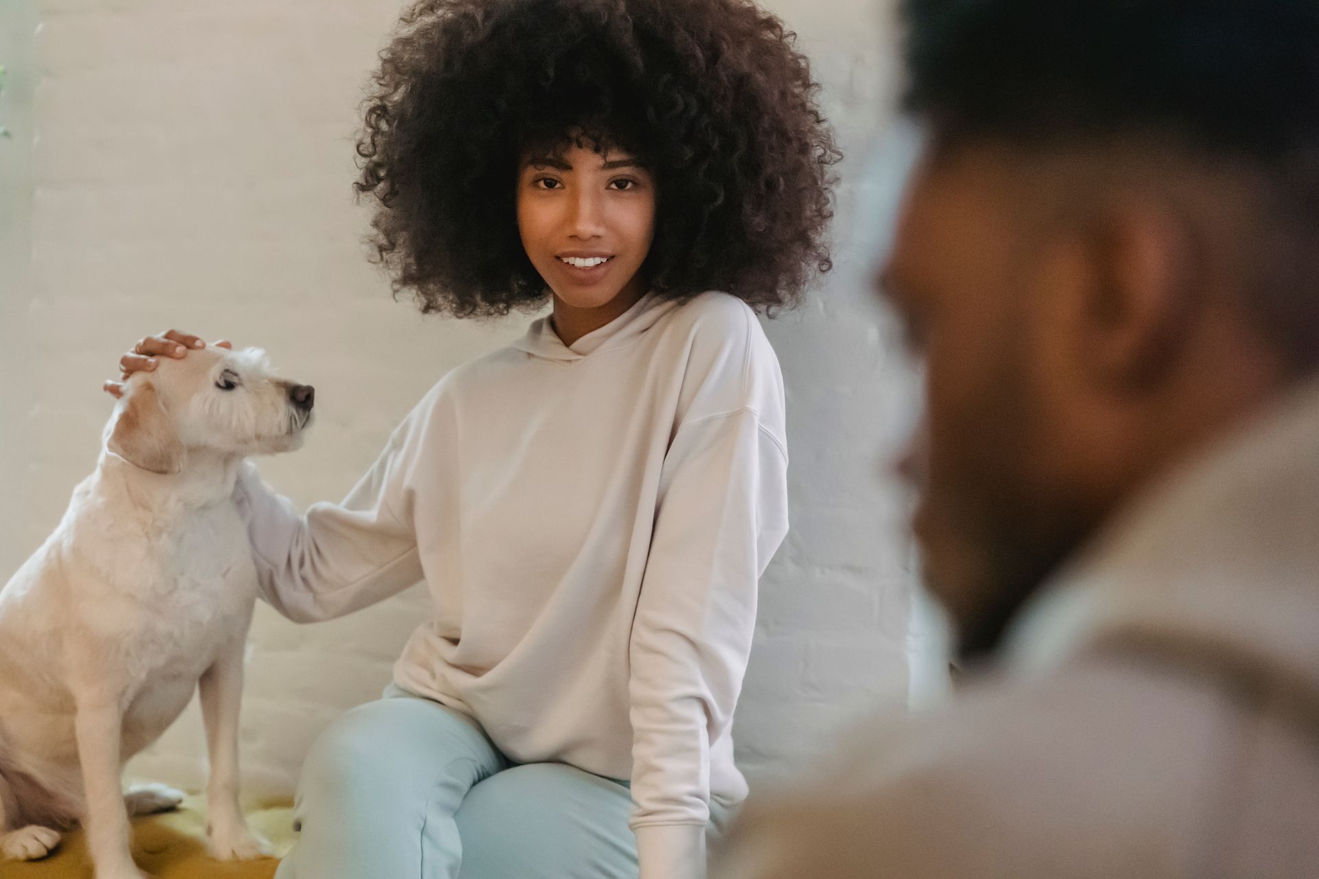 Crop delighted young black lady with dark Afro hair in stylish hoodie petting cute loyal dog and smiling while sitting on chair near focused boyfriend at home