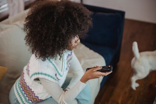 Free From above of focused young black woman with Afro hair in casual outfit sitting on sofa with remote controller in hand and watching TV during weekend at home with dog Stock Photo