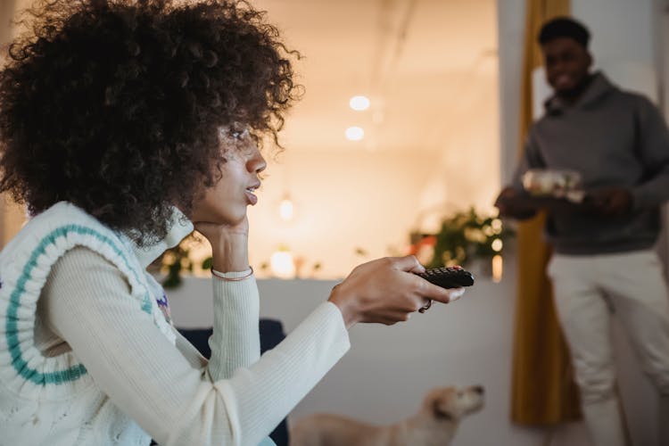 Young Black Woman Watching TV With Interest Near Boyfriend Standing In Room