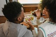 Happy young ethnic woman feeding boyfriend with salad while sitting on couch