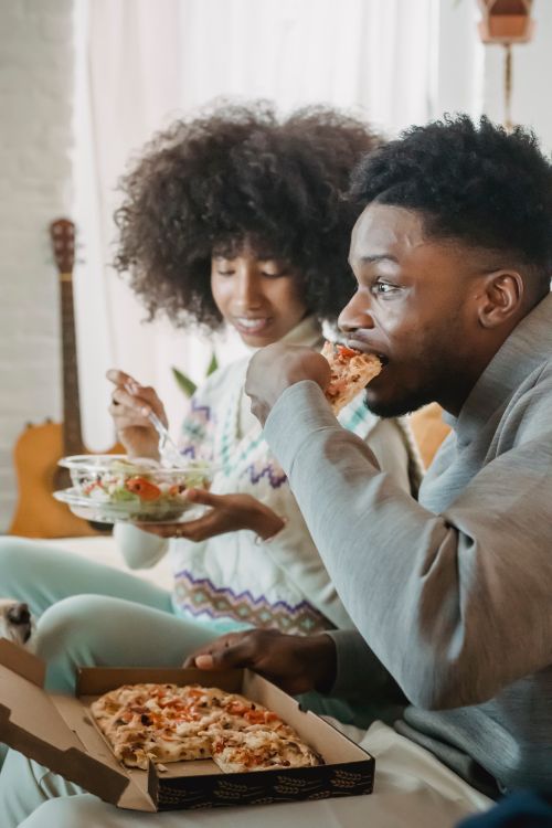 Free Surprised African American crop male eating pizza while ethnic girlfriend having snack with salad Stock Photo