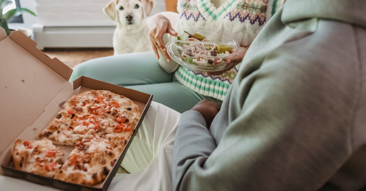 A couple enjoys a cozy meal with pizza and salad while their dog looks on lovingly.