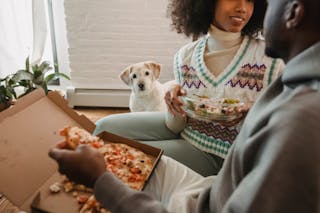 Crop anonymous happy African American couple enjoying delicious salad and pizza while funny cute dog watching