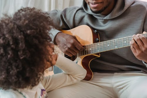 Free Unrecognizable African American girlfriend sitting near talented black boyfriend playing song on guitar while spending time together in light room Stock Photo