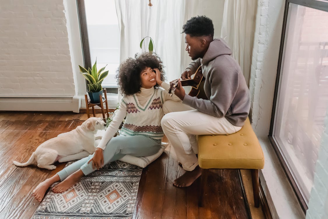 Black man playing guitar for girlfriend in room with dog