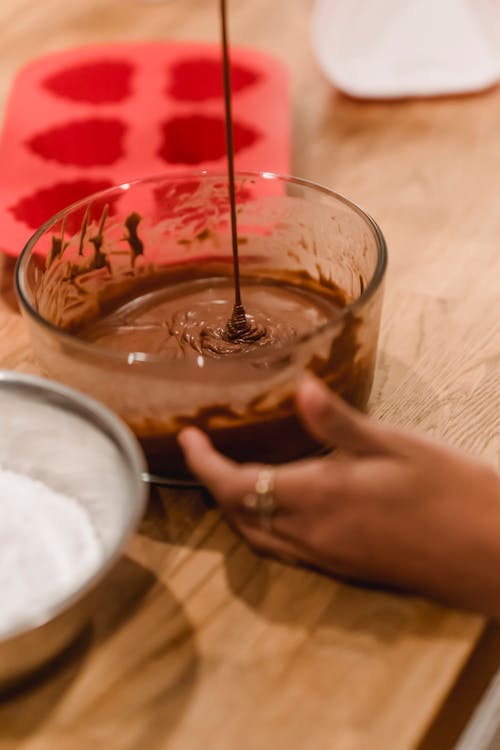 Unrecognizable female cook with glass bowl of chocolate batter for dessert standing at table with silicone molds and flour during cooking