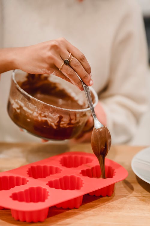 Unrecognizable female cook pouring chocolate batter from bowl into heart shaped silicone molds while cooking pastry at table in kitchen